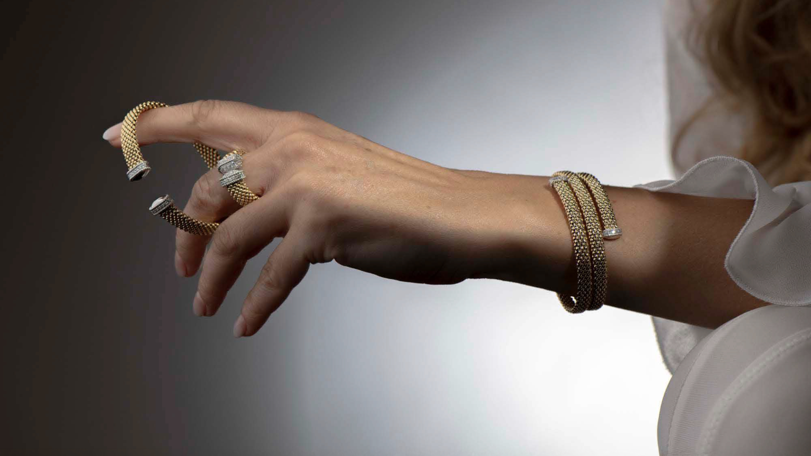 Hand wearing multiple gold bracelets and rings against a gradient background